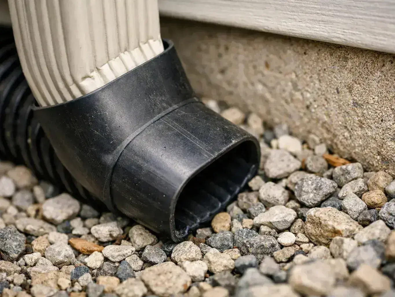 A black downspout elbow rests on gravel beside a concrete wall and white siding
