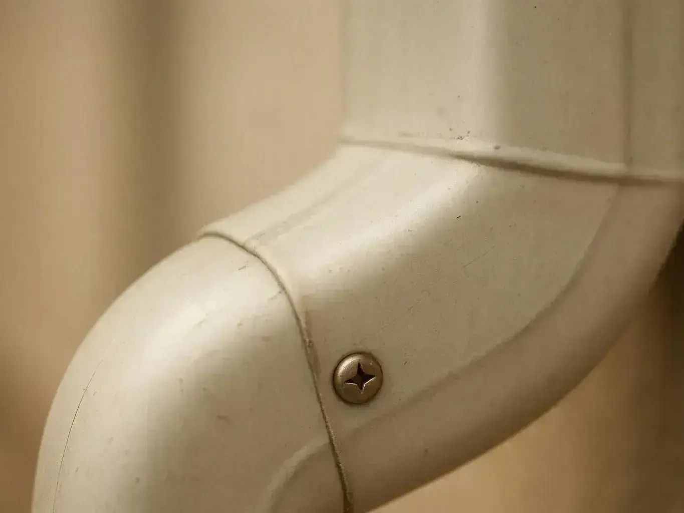 Close-up of a beige plastic pipe elbow with a visible screw against a plain wall