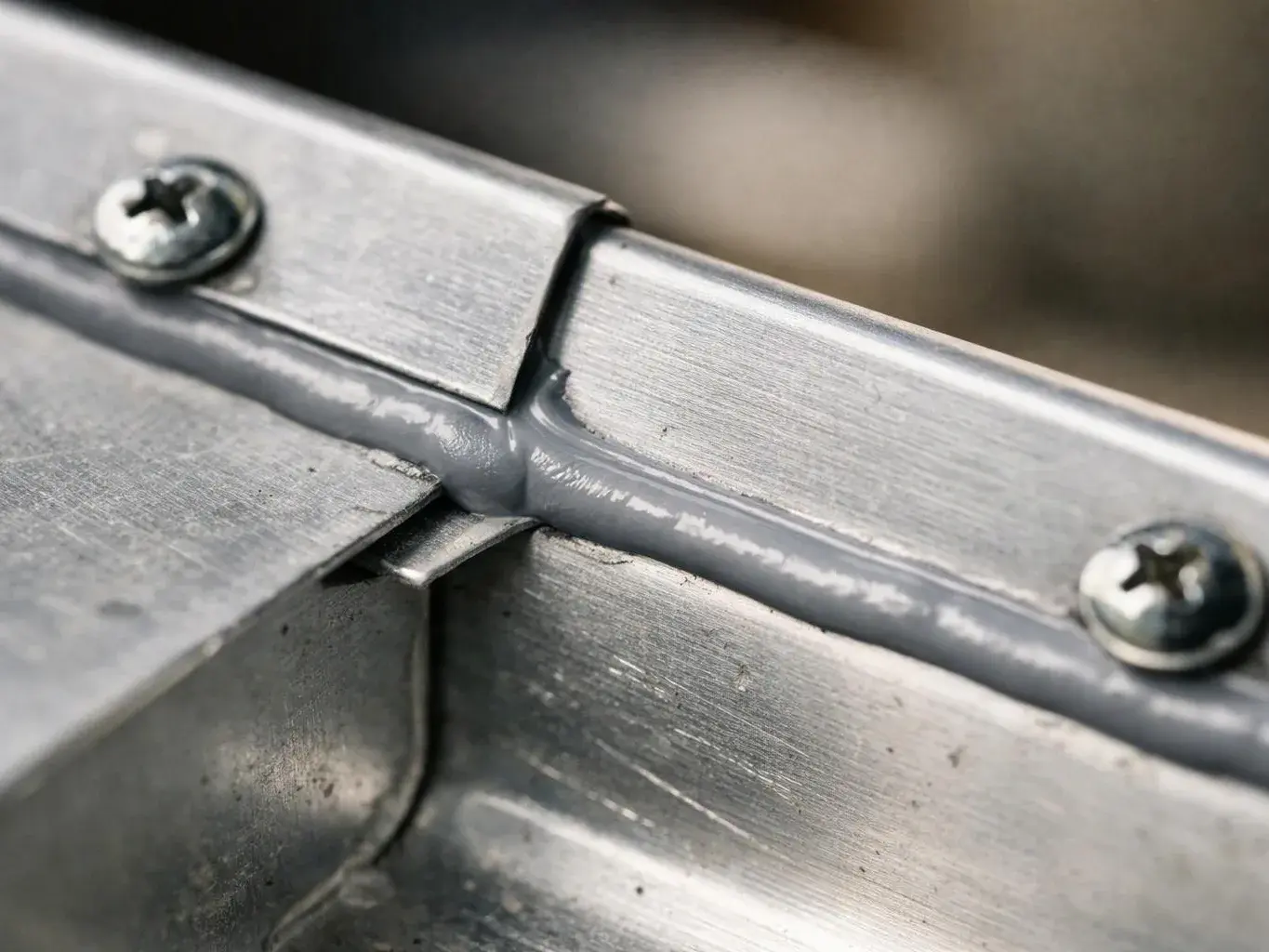 Close-up of brushed metal parts joined by a gray seam, with two screws and a blurred background