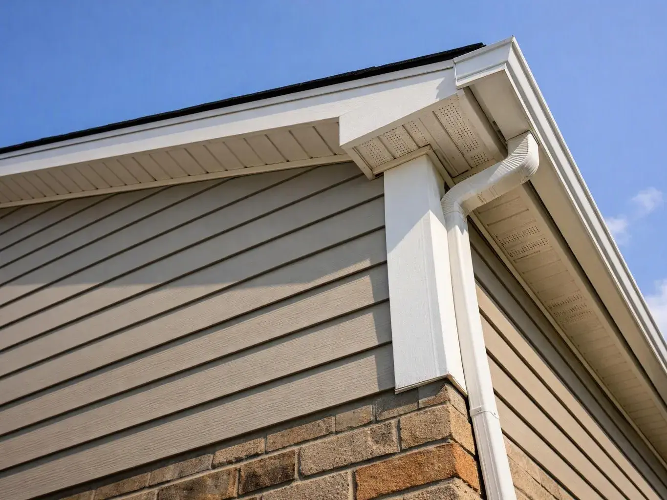 Close-up of a house corner with beige siding, brick below, white trim, and a gutter against blue sky
