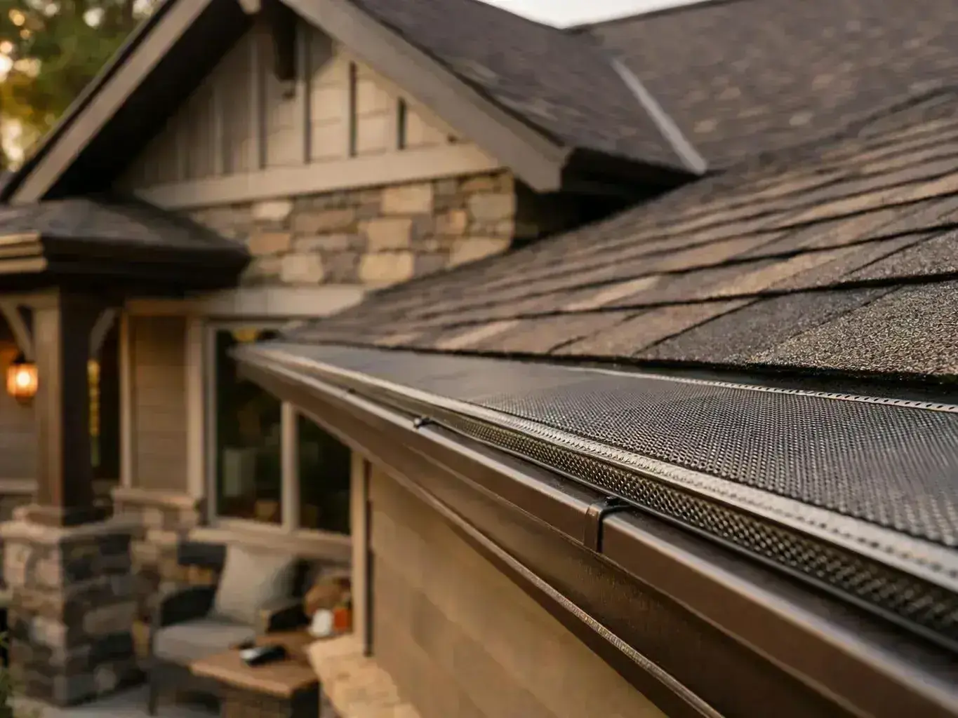 Close-up of a house roof and gutter with leaf guard, with a stone porch and window blurred behind it
