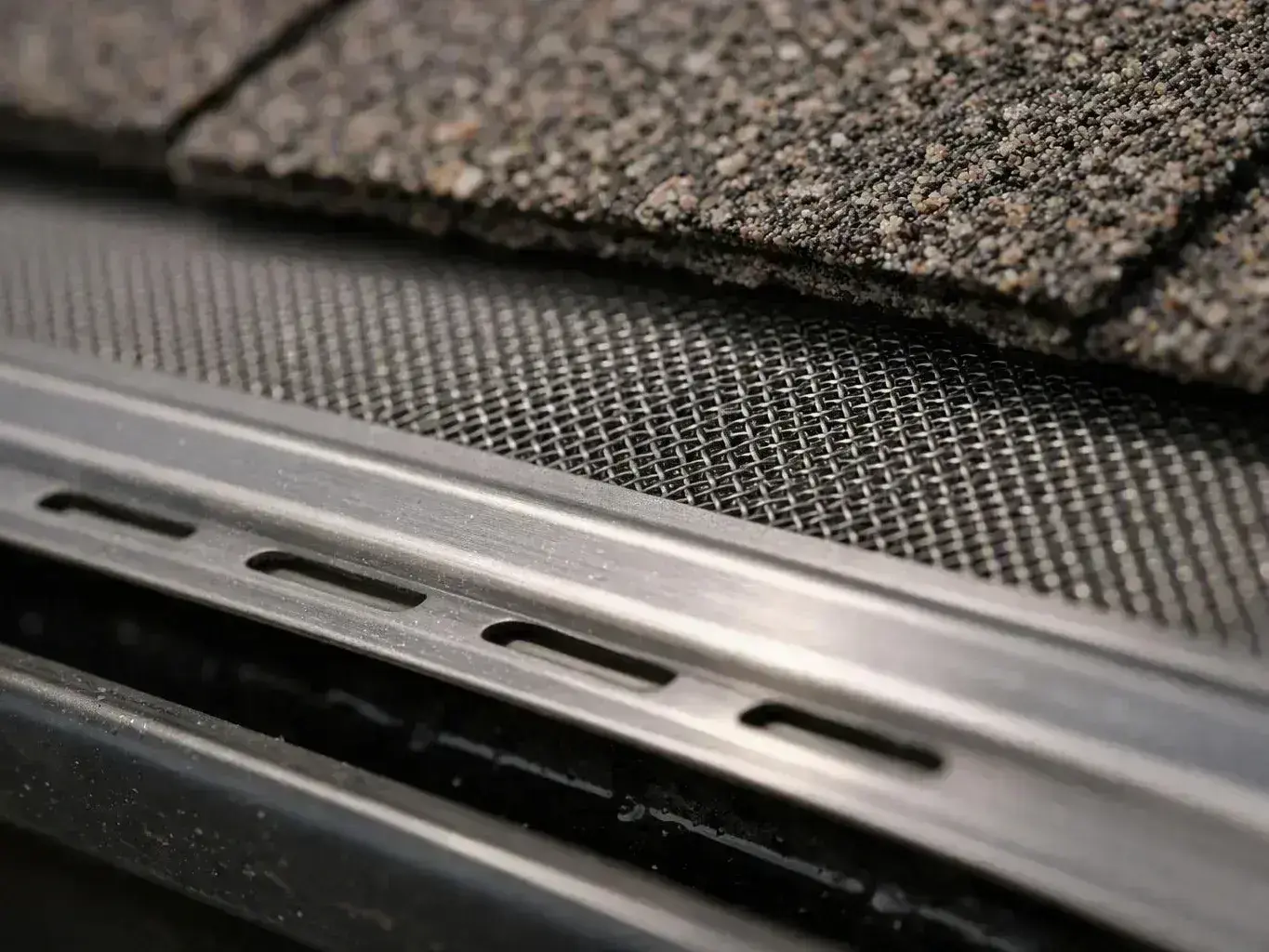 A close-up of layered roofing materials shows granulated shingles, metal mesh, and a slotted metal edge