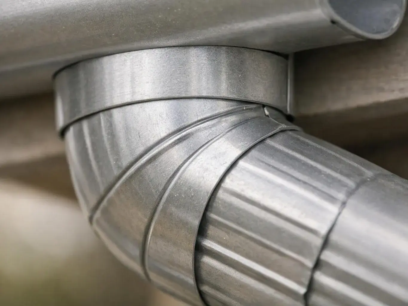 Close-up of a silver metal duct elbow joined to a larger pipe in a plain indoor setting