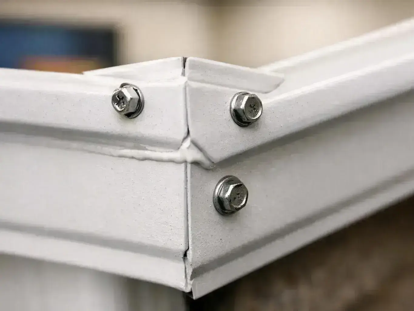 Close-up of a white metal corner bracket with three shiny bolts against a blurred indoor background
