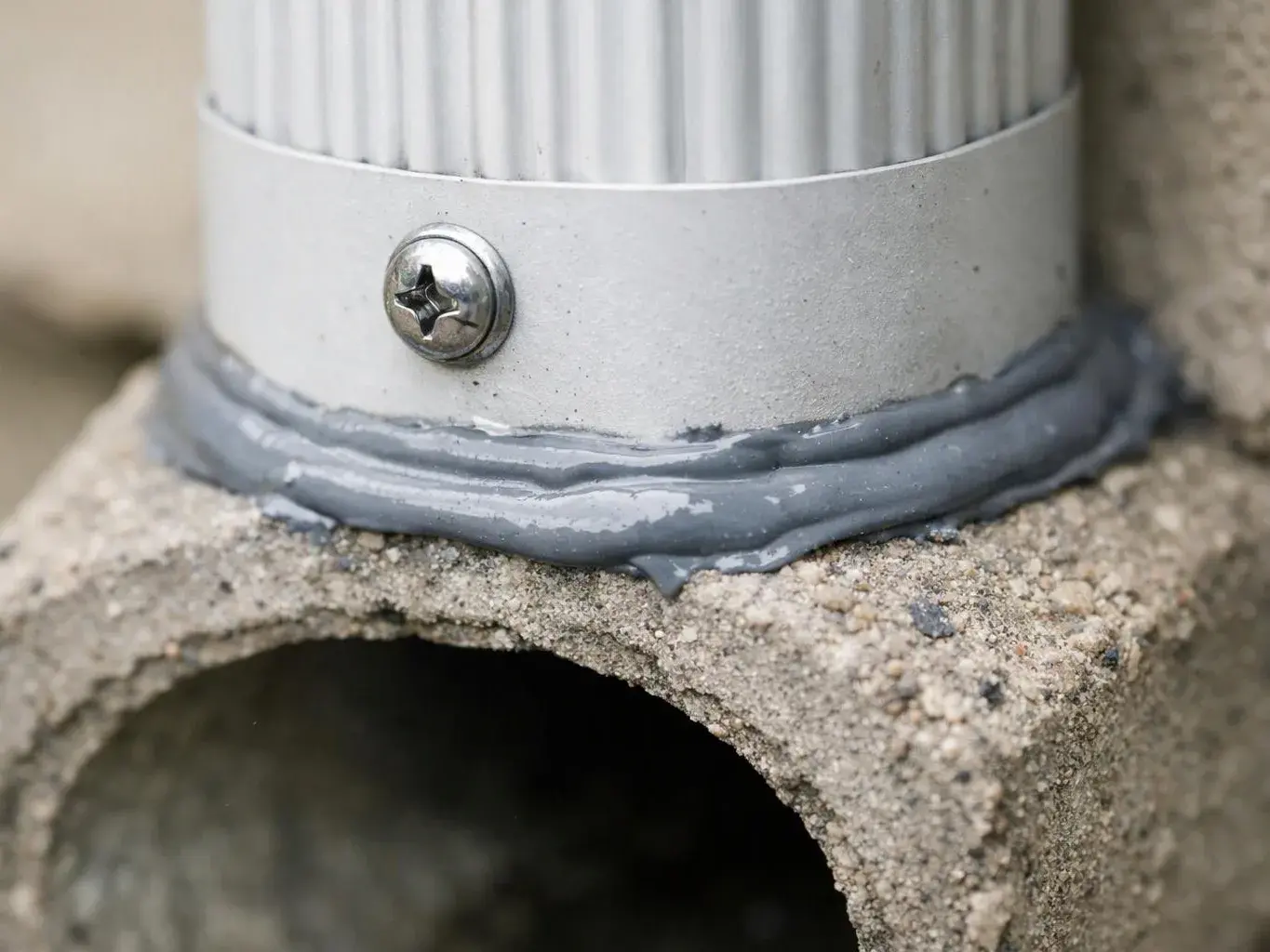 Close-up of a white ribbed pipe secured with a screw and sealed to rough concrete with gray caulk