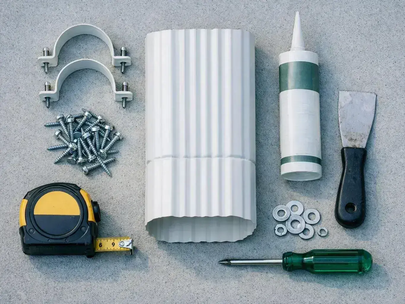 A flat lay of gutters, screws, clamps, sealant, a putty knife, tape measure, washers, and screwdriver on gray concrete