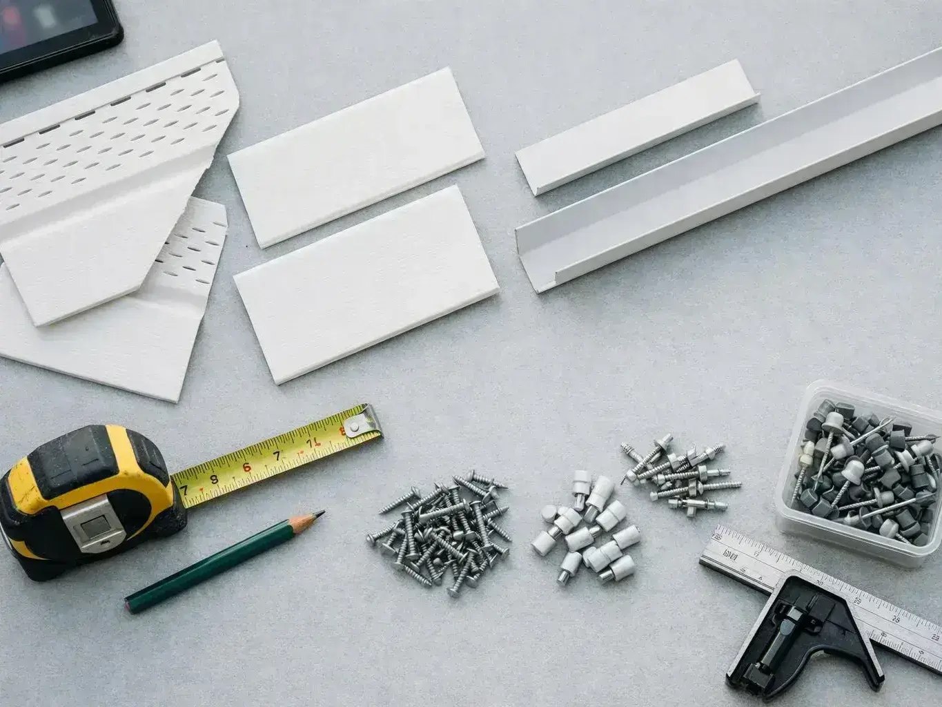 Flat lay of white trim pieces, screws, anchors, a tape measure, pencil, ruler, and a stapler on a gray surface