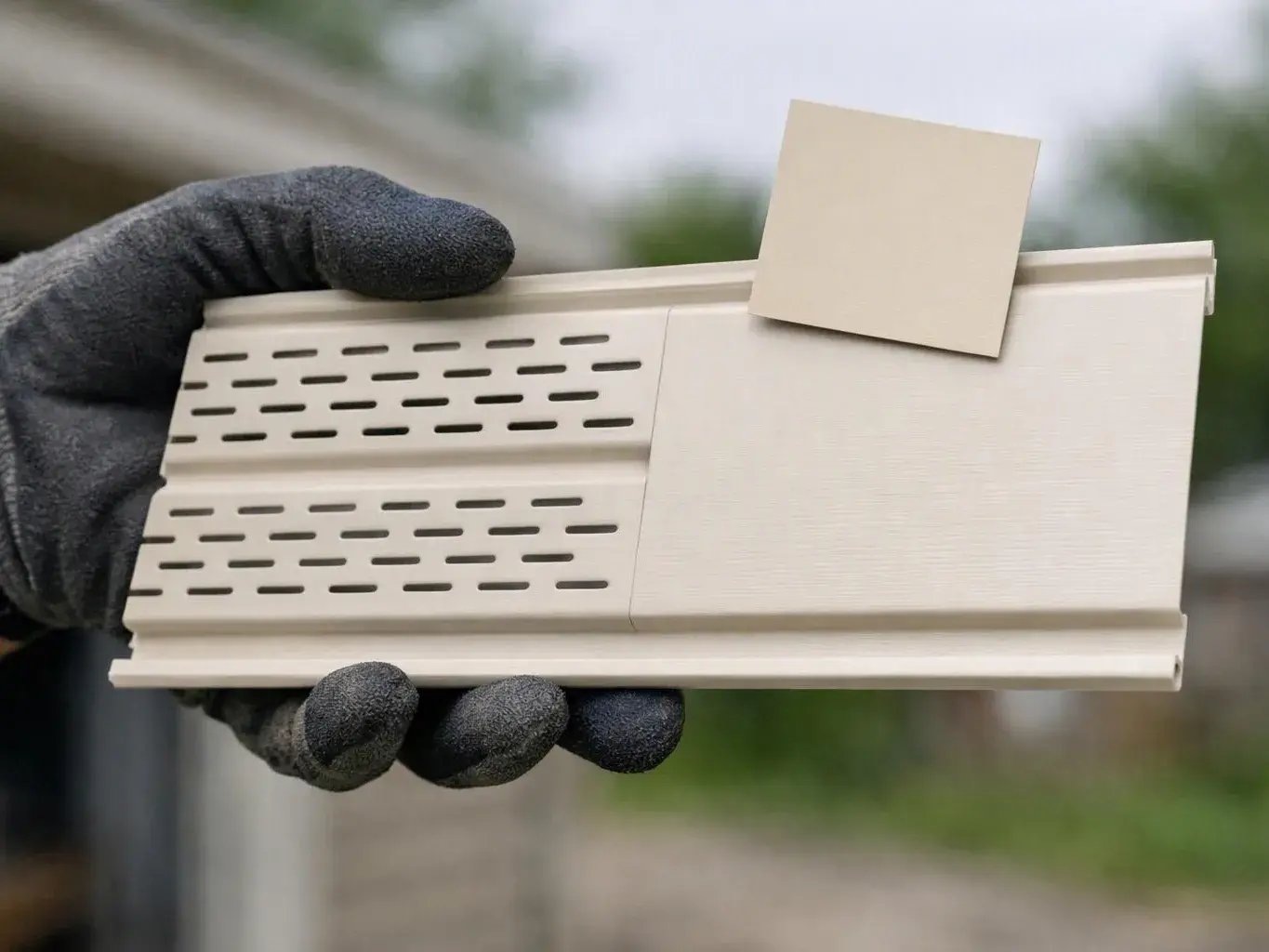 A gloved hand holds a cream vented siding sample outdoors with a small color swatch attached