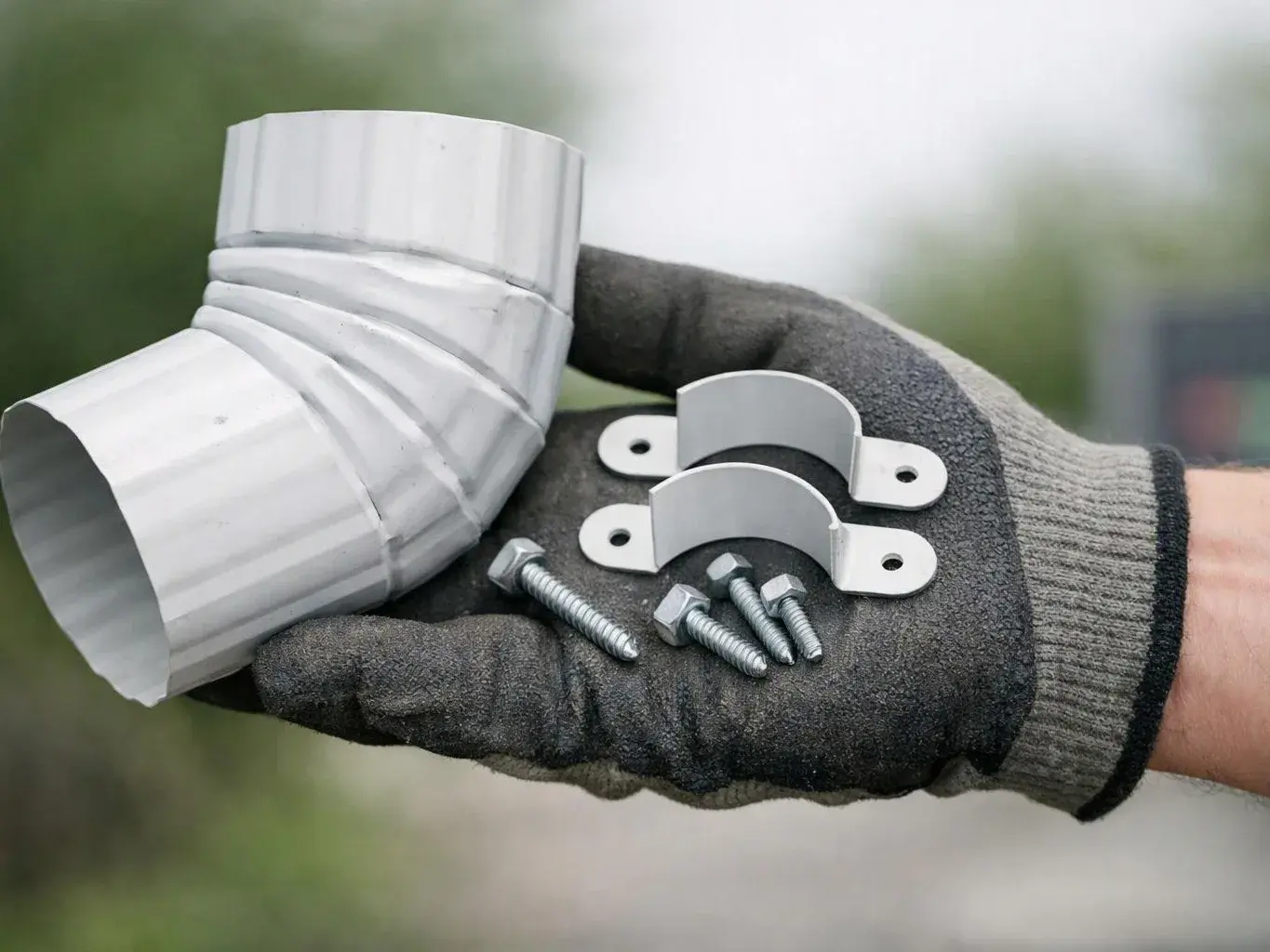 A gloved hand holds a white metal elbow pipe fitting, two clamps, and several screws outdoors