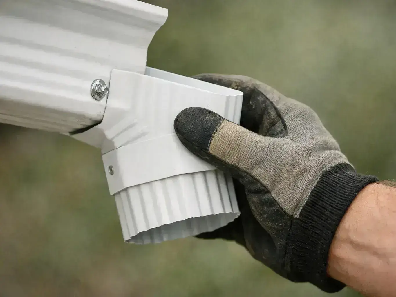 A gloved hand holds a white metal gutter corner with a downspout elbow against a blurred green background