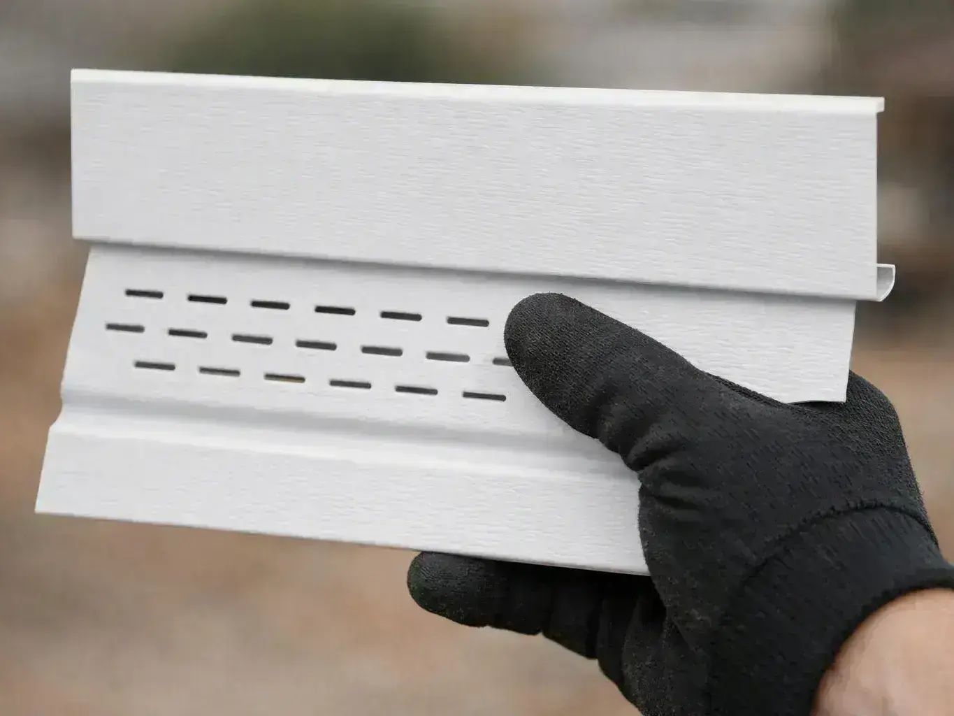 A gloved hand holds a white vinyl siding sample with rows of small black slots outdoors