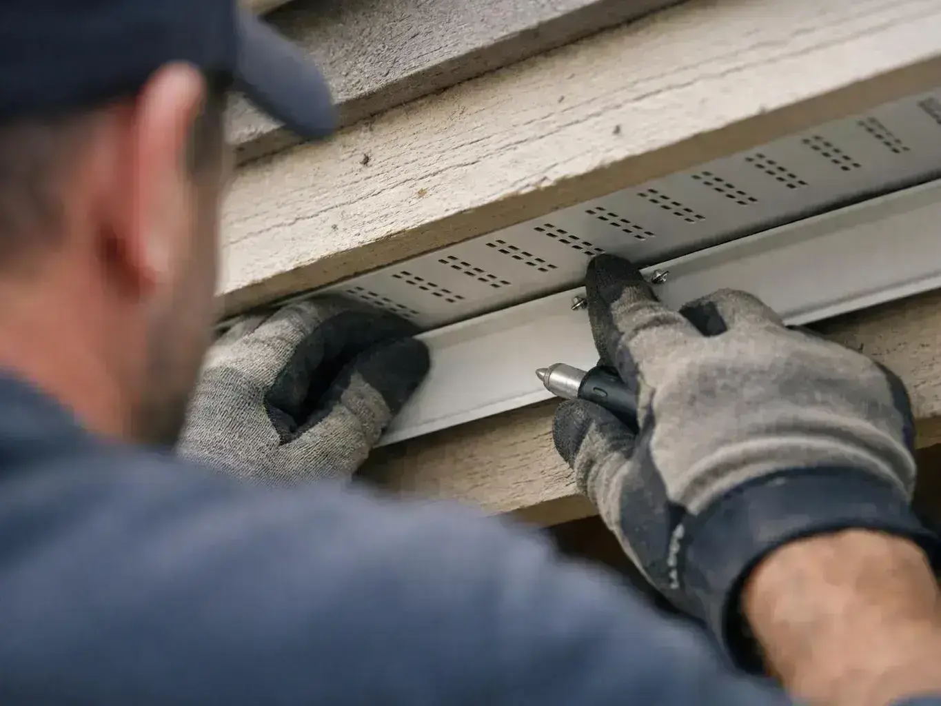 A gloved worker fits a white metal strip under house siding with a handheld tool