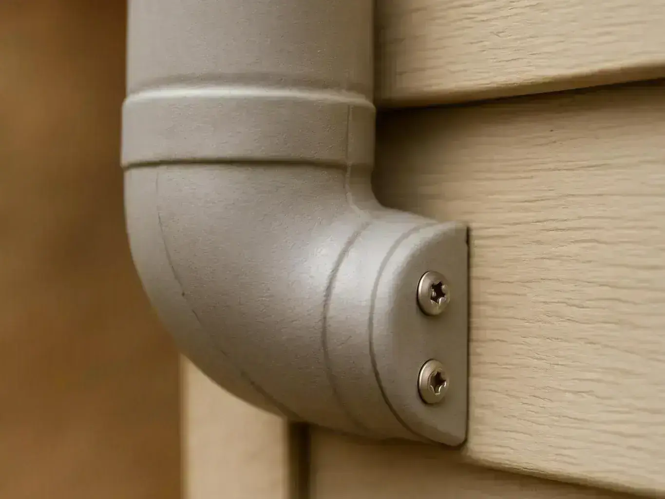 A gray metal elbow pipe is screwed to beige wooden siding in close-up