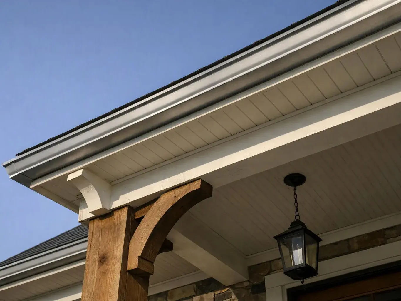 A house roof eave with white trim, wooden support beams, and a hanging black lantern against a blue sky