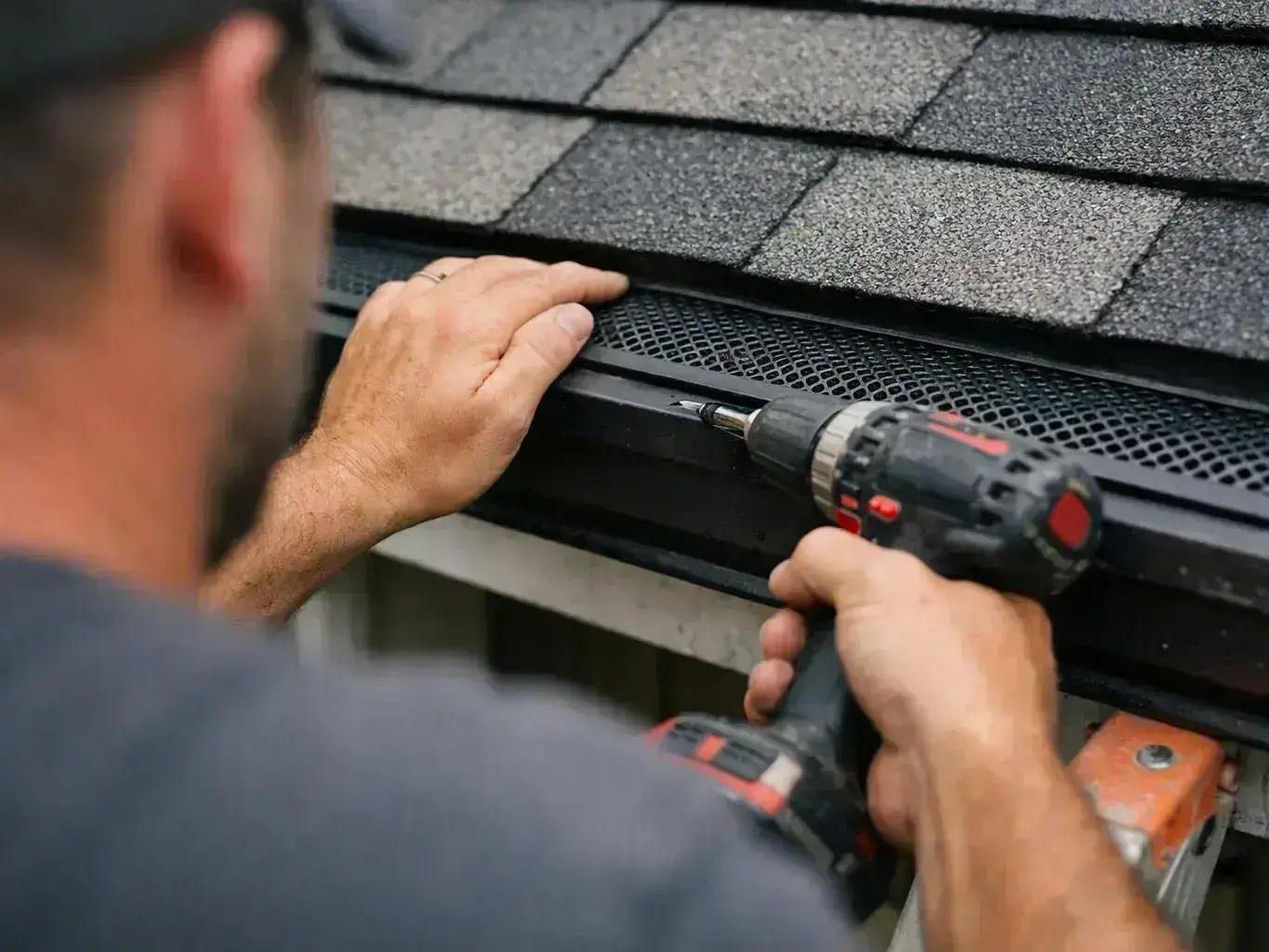 A man uses a cordless drill to fasten trim along a shingled roof edge