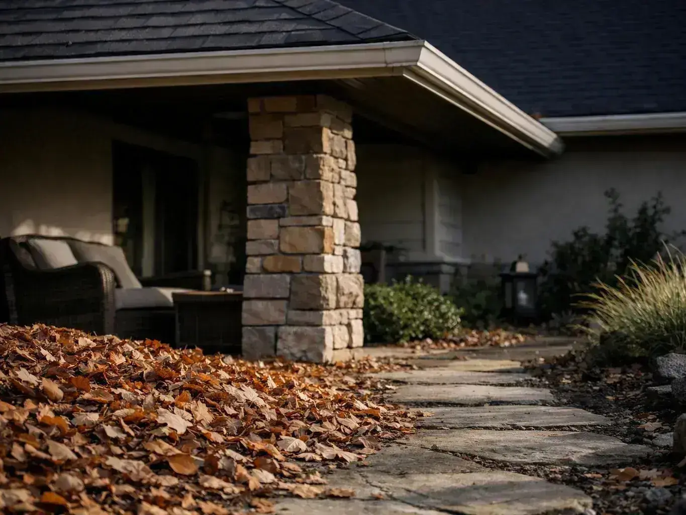 A stone walkway winds past a porch covered with fallen autumn leaves outside a house