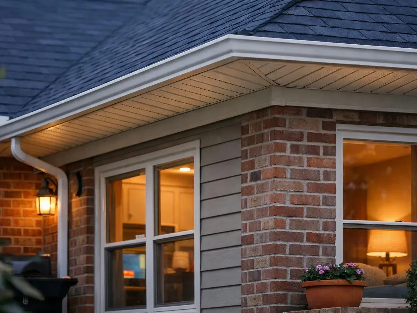 Warm porch lights glow on a brick and siding house with windows, a flower pot, and a grill under the roofline