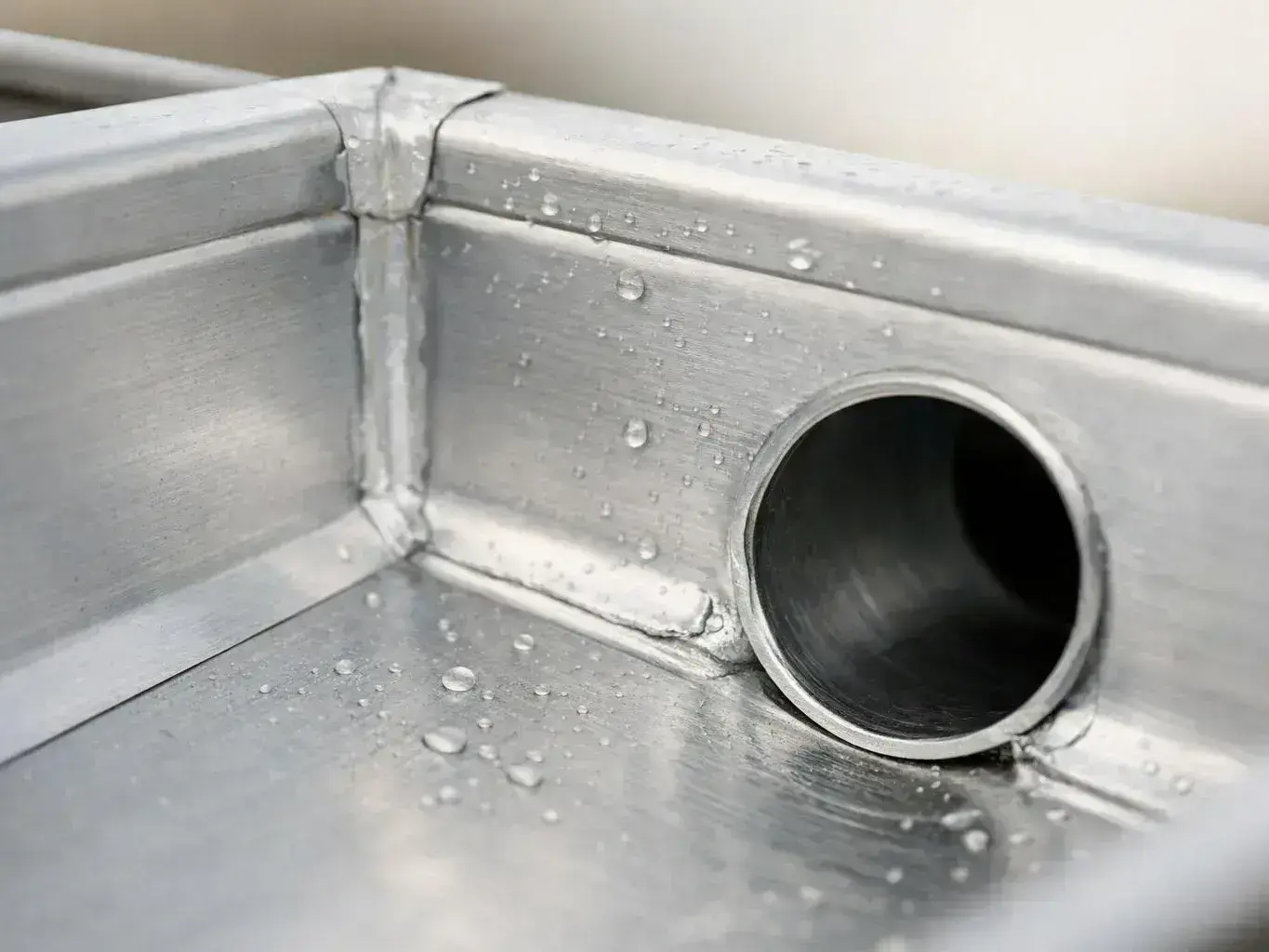 Water droplets sit on a metal sink corner beside a round drain opening