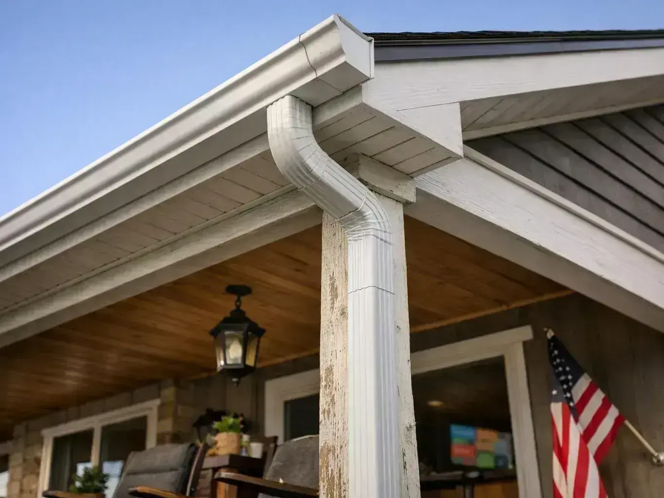 A white downspout runs from the roof gutter beside a porch with a hanging lantern and an American flag