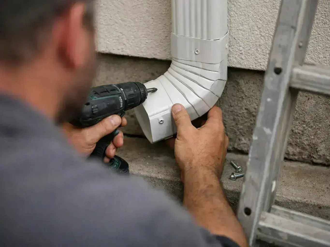 A worker drills screws into a white downspout elbow beside a stucco wall and ladder