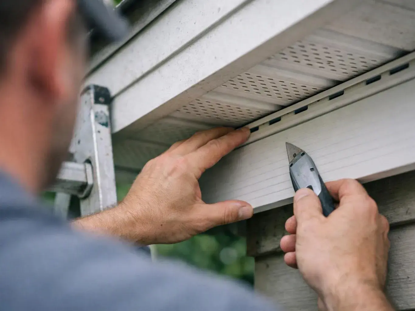 A worker fits a white trim board under a roof edge with a utility knife outdoors